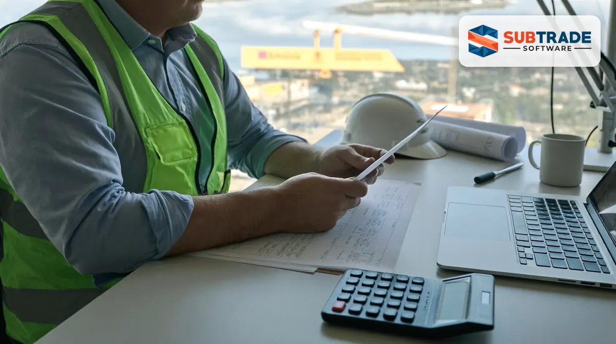 A subcontractor reviewing payment and holdback figures on a laptop at a site office desk with a calculator and construction drawings beside it.